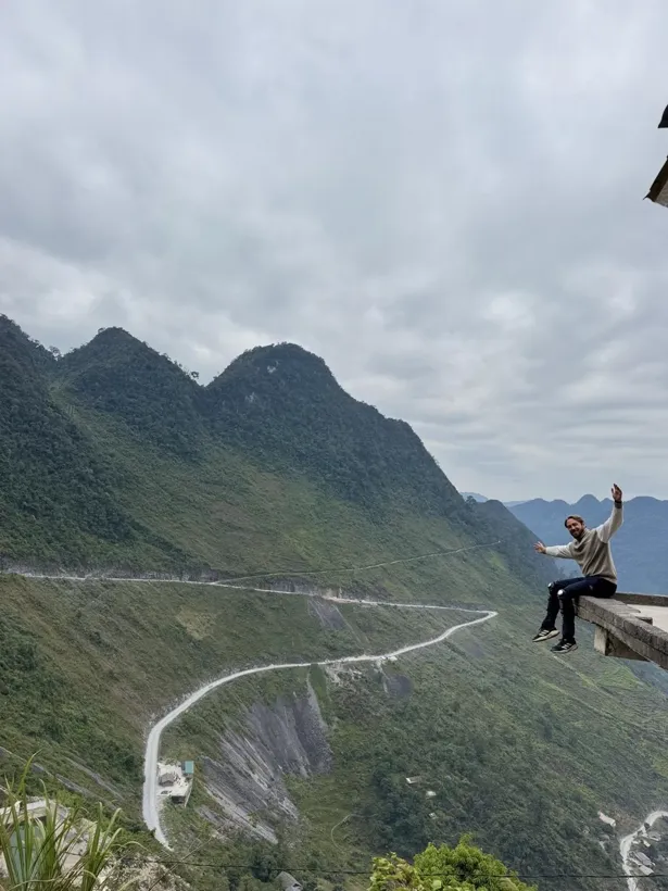 Brent zit op de rand van een uitkijkpunt en zwaait, boven een slingerende bergweg